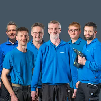 Montageteam von Möbel König in blauer Arbeitskleidung mit Werkzeug vor grauem Hintergrund.